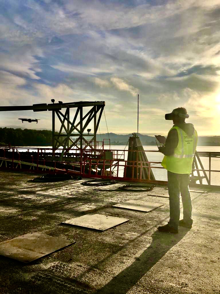 Mark Gibson in a high-visibility vest and FPV goggles, tablet in hand, flying a maritime inspection with a drone airborne over the water beside industrial deck structures