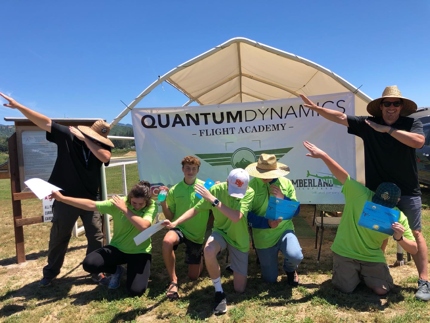 Quantum Dynamics Flight Academy youth STEM graduates in green academy shirts holding their certificates in front of the academy banner, celebrating with instructor Sean Holt