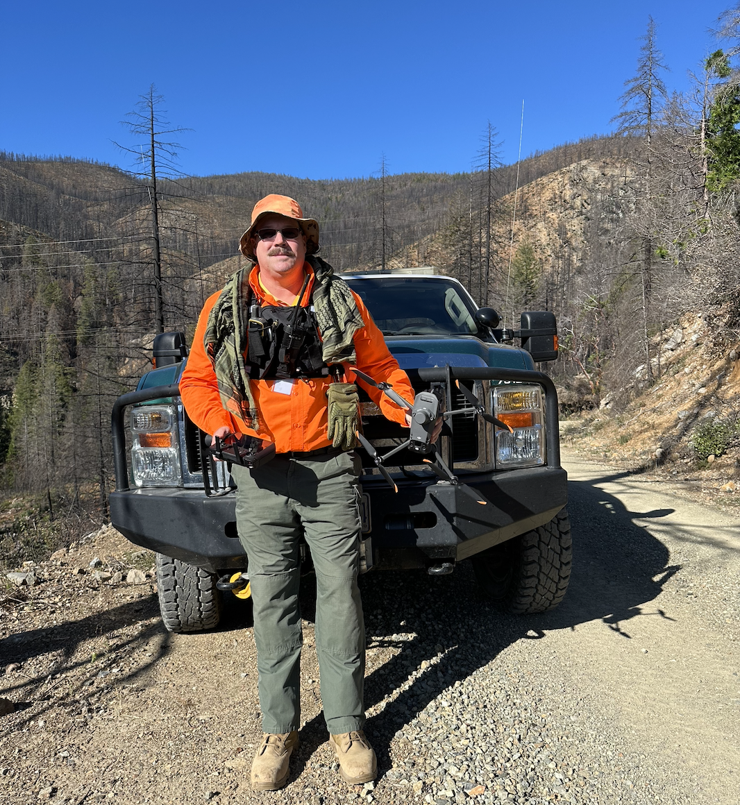 Sean Holt in orange search and rescue gear with a radio chest rig, holding a drone and controller in front of a truck in burned wildfire country